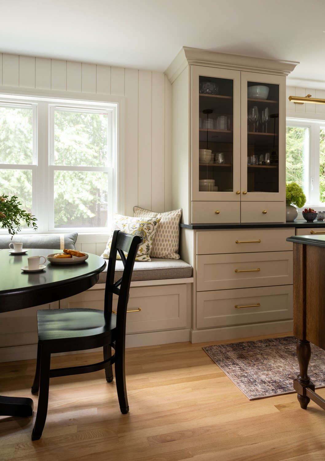 Built-in window seat with a round table and a black chair beside tall glass-front cabinets