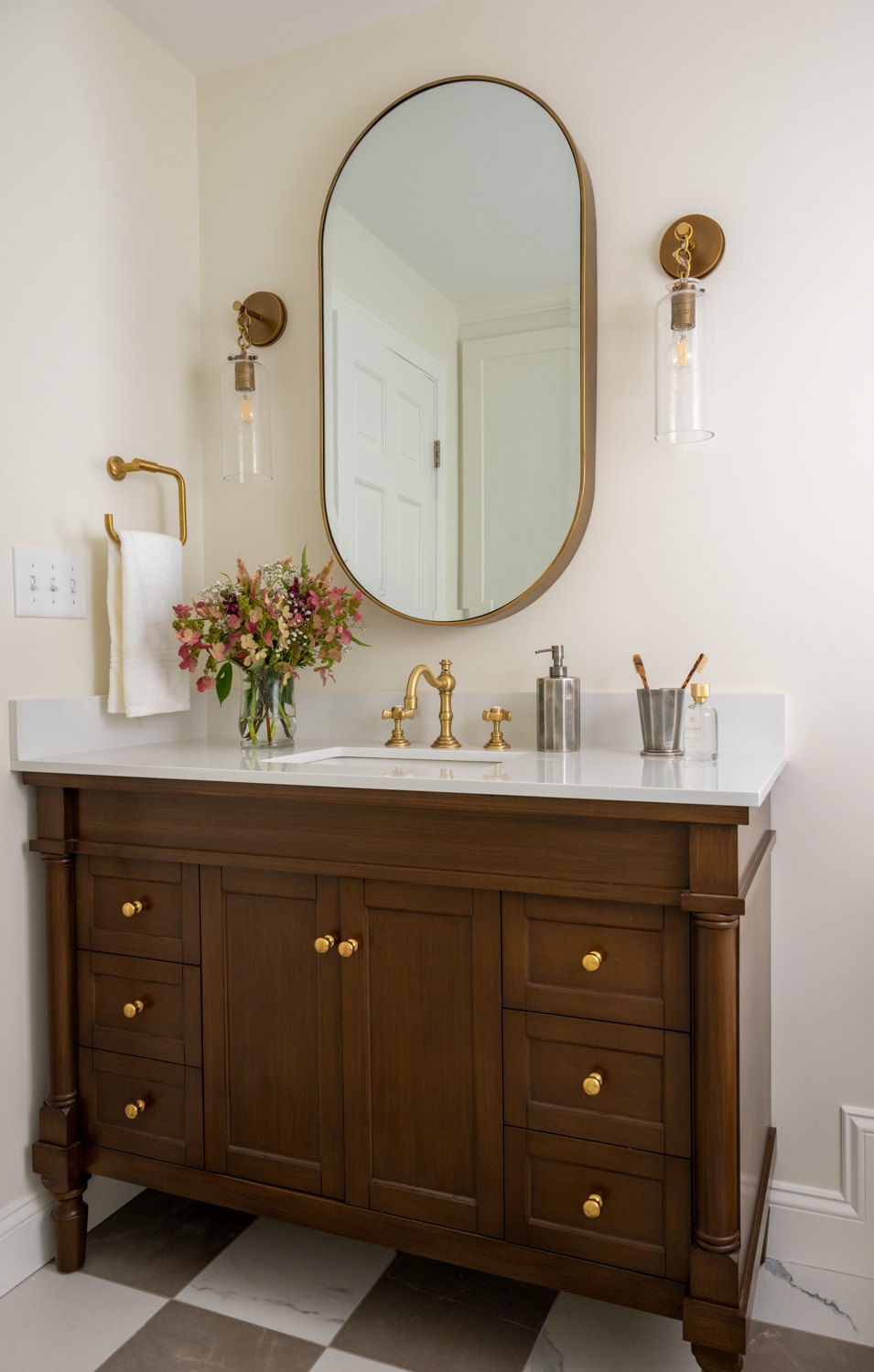 Cherry vanity with an oval mirror and paired sconces on a checkerboard floor